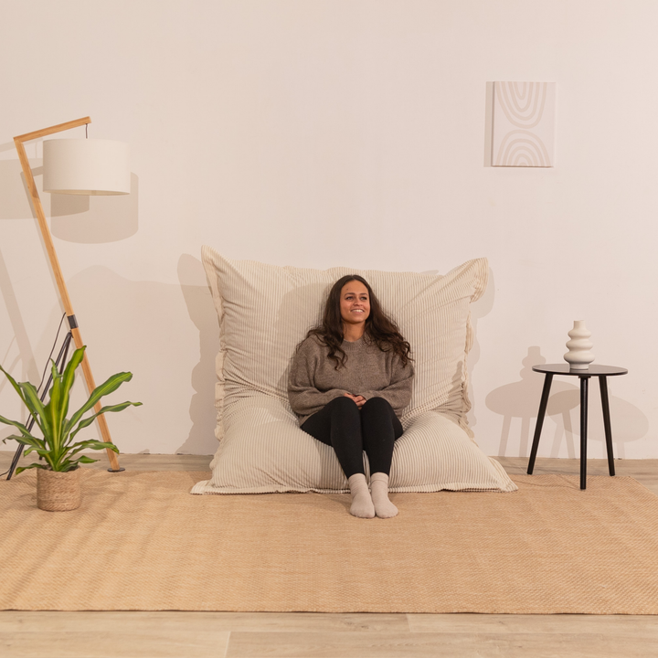 Personne assise sur un pouf 140x180 cm blanc cassé en velours côtelé, dossier appuyé contre le mur dans un décor d&