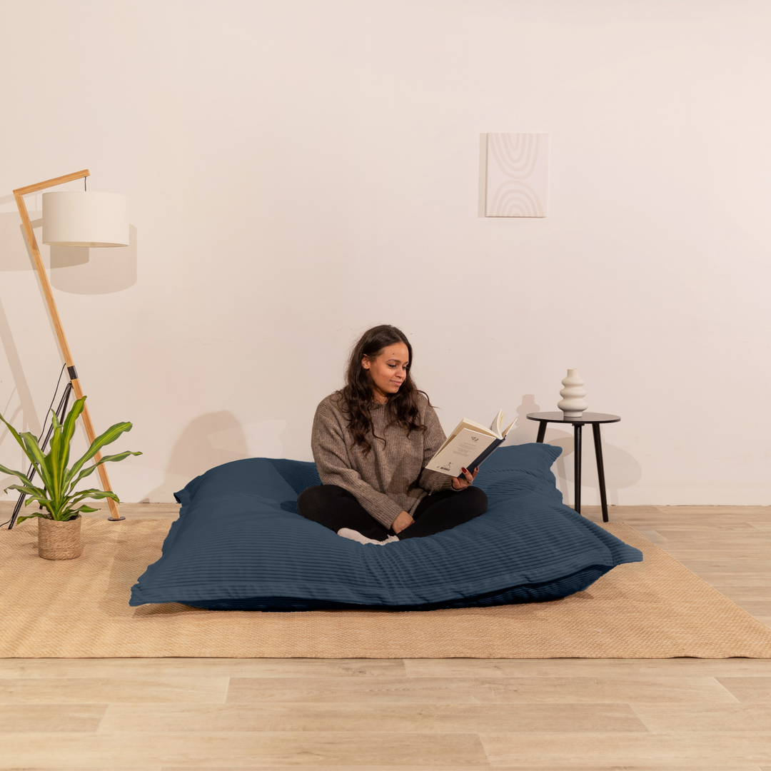 Personne assise sur un pouf 140x180 cm bleu nuit en velours côtelé, dans un décor d&
