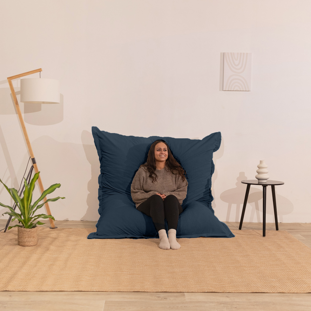 Personne assise sur un pouf 140x180 cm bleu foncé en velours côtelé, dossier appuyé contre le mur dans un décor d&