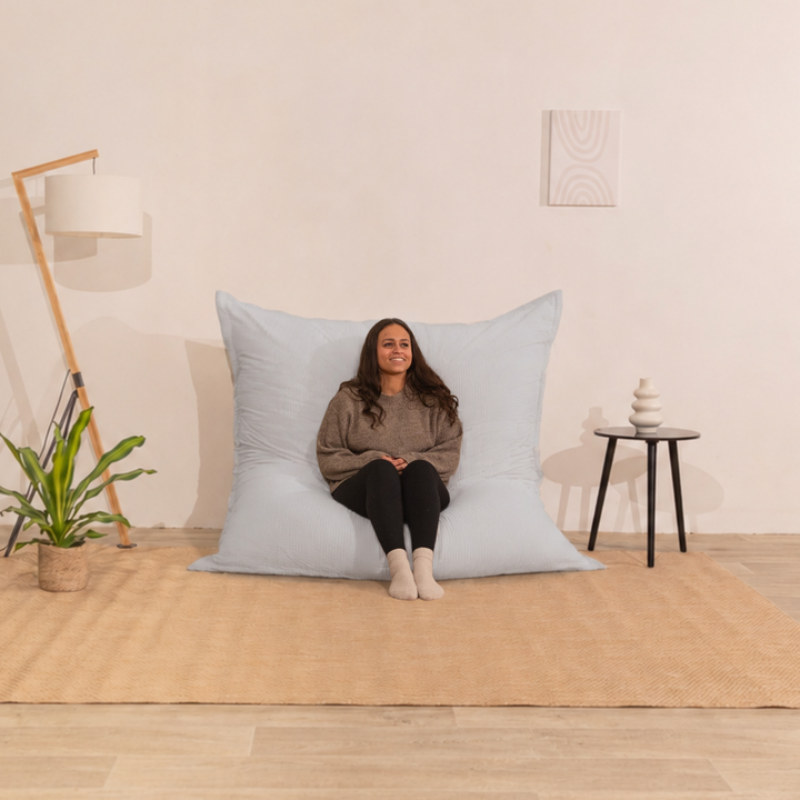 Personne assise sur un pouf 140x180 cm gris clair en velours côtelé, dossier appuyé contre le mur dans un décor d&